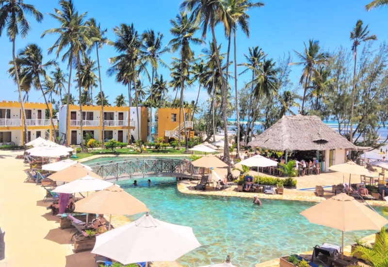 Resort pool with lounge chairs and umbrellas, tropical palm trees, beachfront bar, and ocean view on a sunny day
