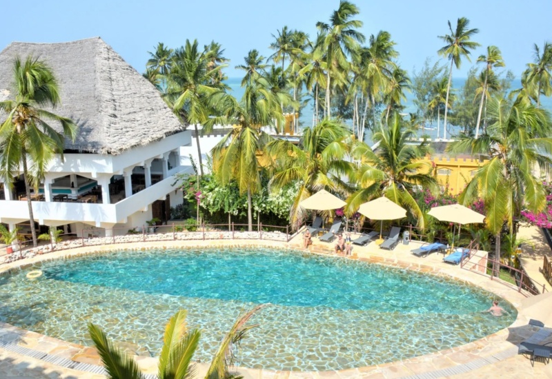 Resort pool with clear water surrounded by lounge chairs, palm trees, thatched-roof dining pavilion, and ocean view in the background