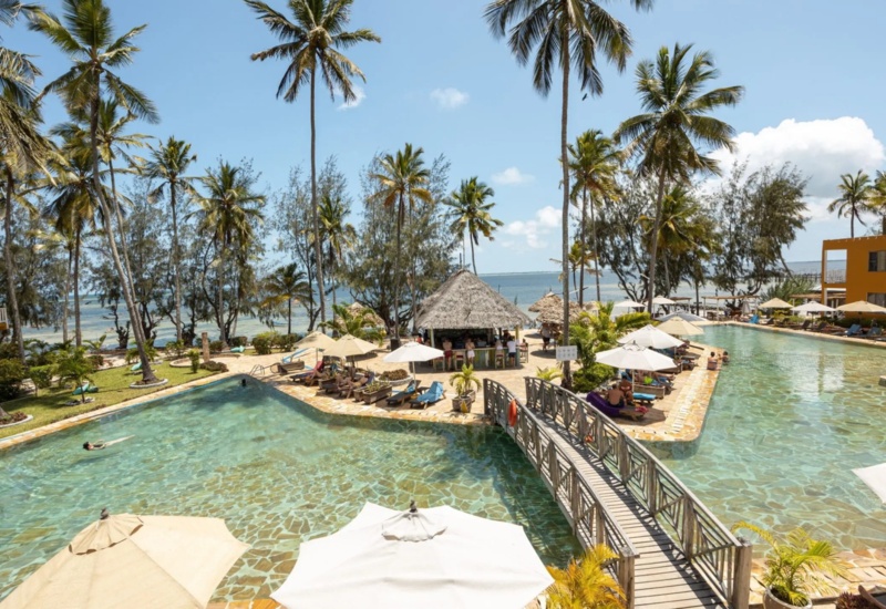 Resort pool area with two large pools separated by a wooden bridge, poolside bar under a thatched roof, palm trees, and ocean view