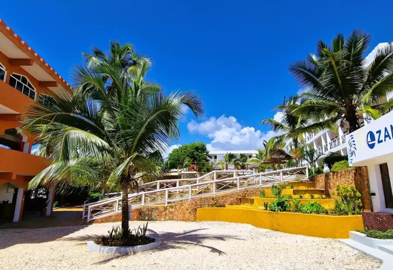 Bright tropical hotel courtyard with palm trees, colorful buildings, stone pathways, and clear blue sky
