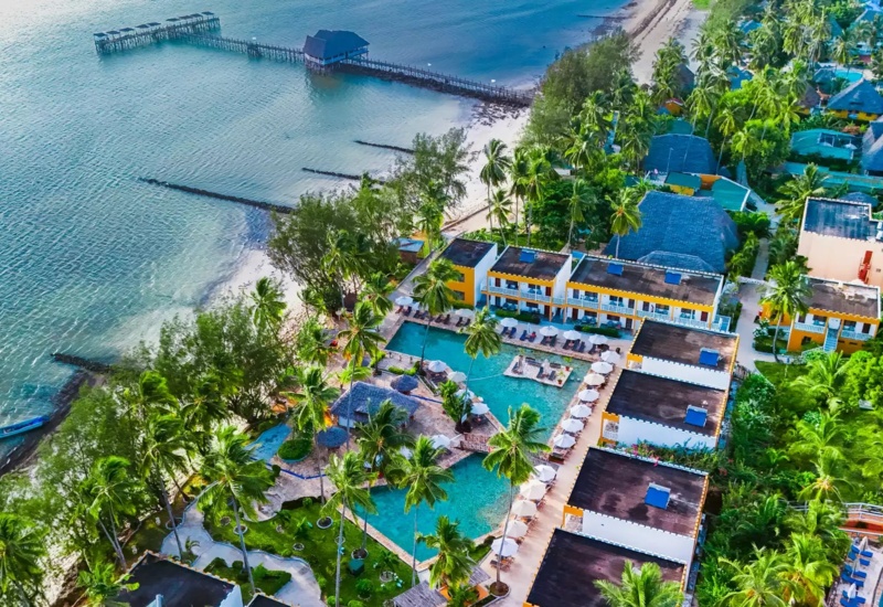 Beachfront resort with pool surrounded by palm trees, sun loungers, and a wooden pier extending into calm ocean waters