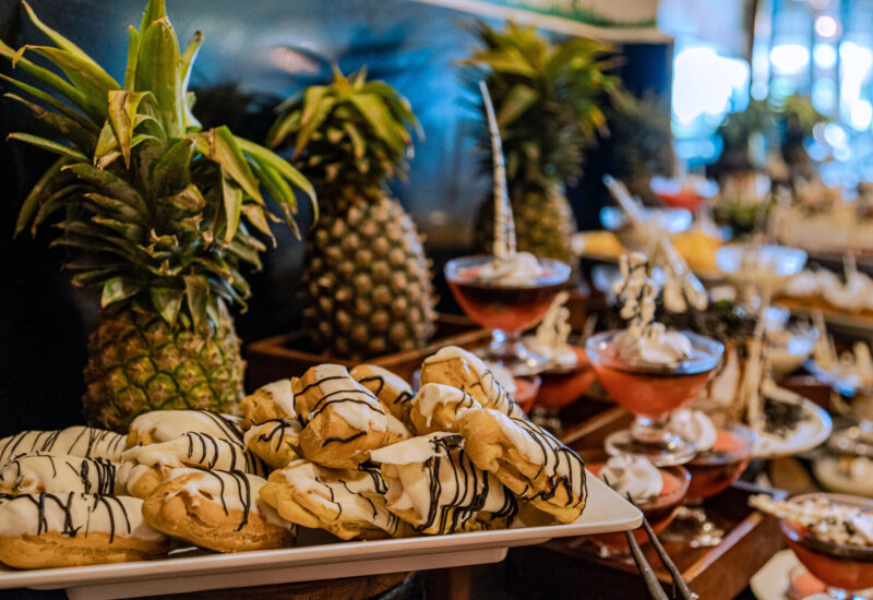 Tray of cream-filled eclairs with white and dark chocolate drizzle beside fresh pineapples and layered dessert glasses in buffet setting