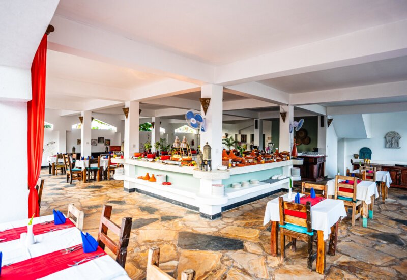 Bright hotel dining area with rustic wooden chairs, white tablecloths, red runners, blue napkins, and a central buffet counter