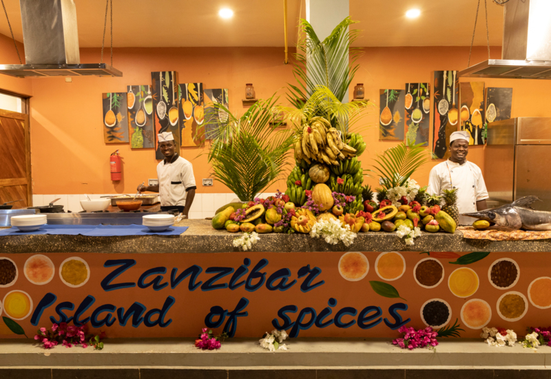 Two chefs smiling behind a vibrant counter decorated with fresh tropical fruits, palm leaves, and a large fish sculpture in a warm kitchen