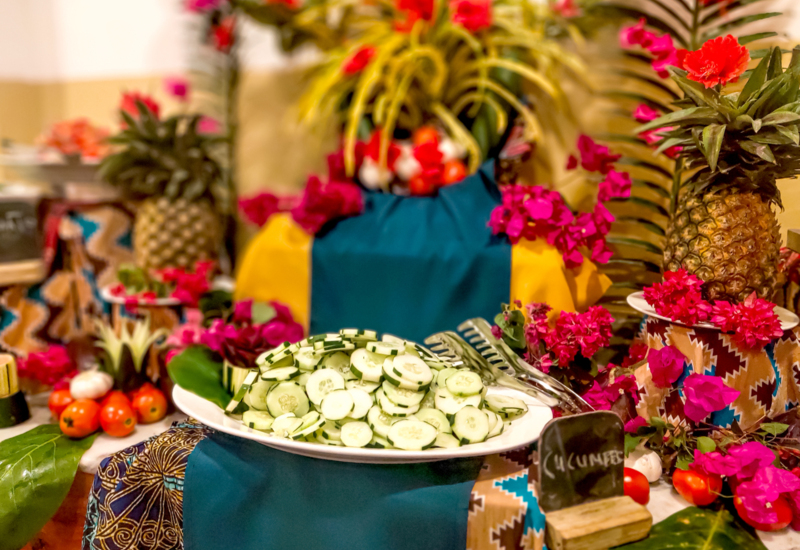 Fresh cucumber slices on a white plate surrounded by tropical decor with pineapples, tomatoes, and vibrant pink flowers