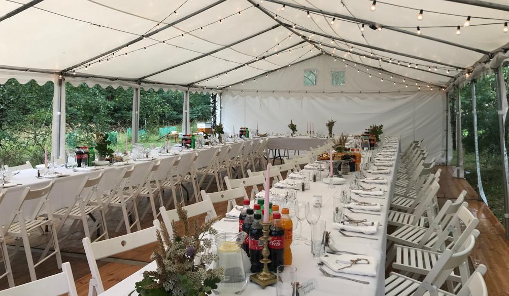Przyjęcie w dużym namiocie z długimi stołami, białymi krzesłami, dekoracjami i napojami, w otoczeniu zieleni