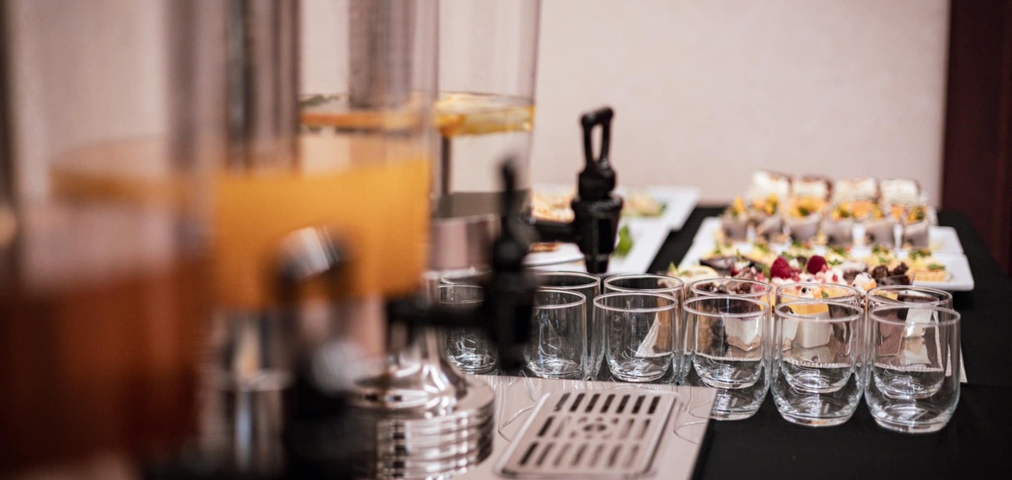 Stół z dystrybutorami świeżych soków i zestawem przeźroczystych szklanek przed bufetem z różnorodnymi deserami i przekąskami