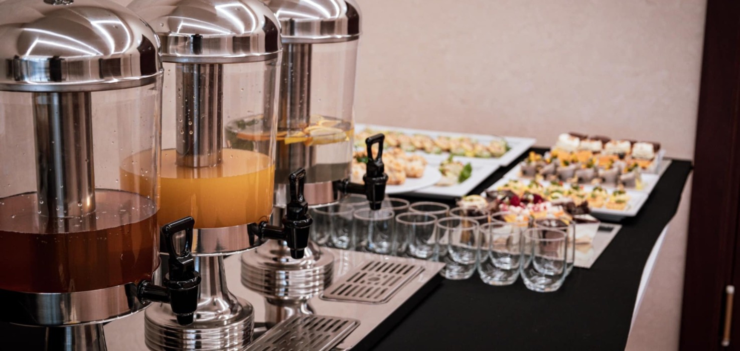 Stół bufetowy z trzema dozownikami napojów i różnorodnym wyborem przekąsek i deserów na białych półmiskach