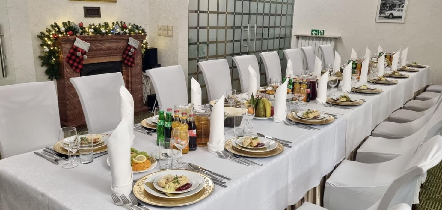 Długi stół bankietowy udekorowany białym obrusem i zastawą, przy kominku z świątecznymi skarpetami i girlandą świetlną.