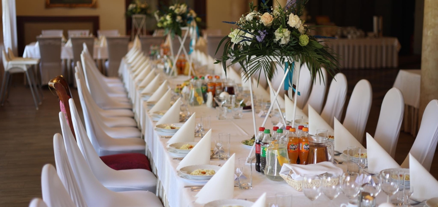 Długi stół bankietowy nakryty białym obrusem, z białymi serwetkami, kwiatowymi dekoracjami i napojami w eleganckiej sali na przyjęcie