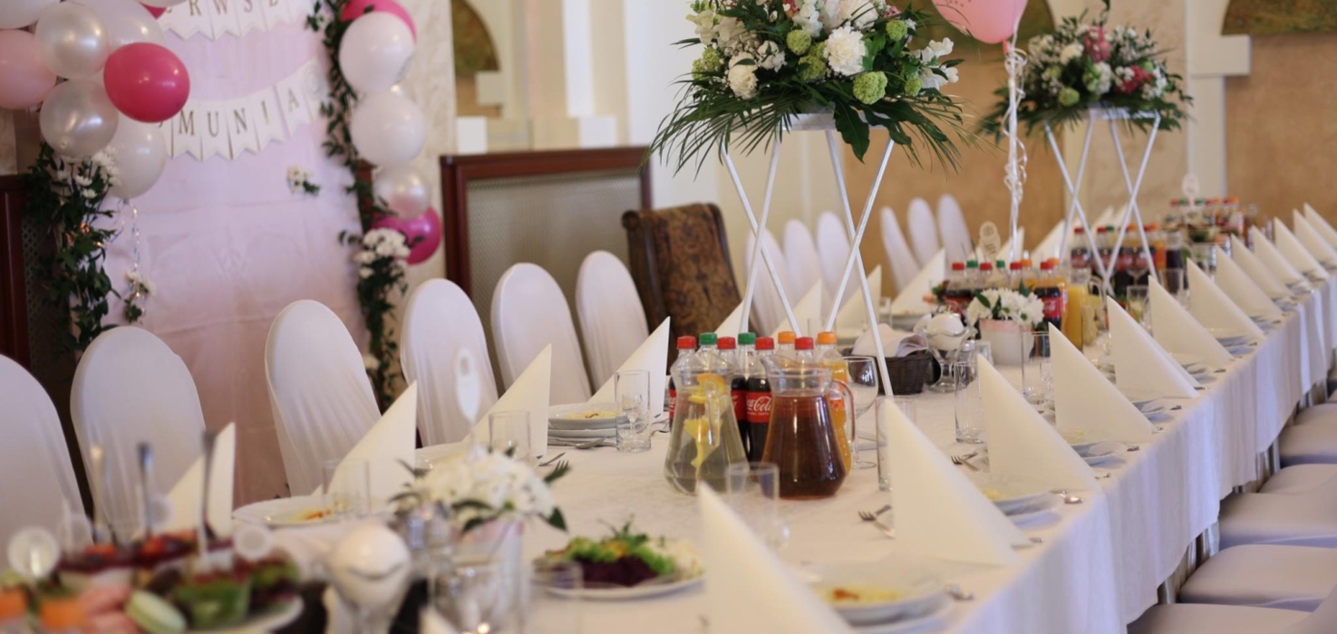 Długa sala bankietowa ozdobiona białymi i różowymi balonami, elegancko nakryty stół z kwiatami i napojami na uroczystość