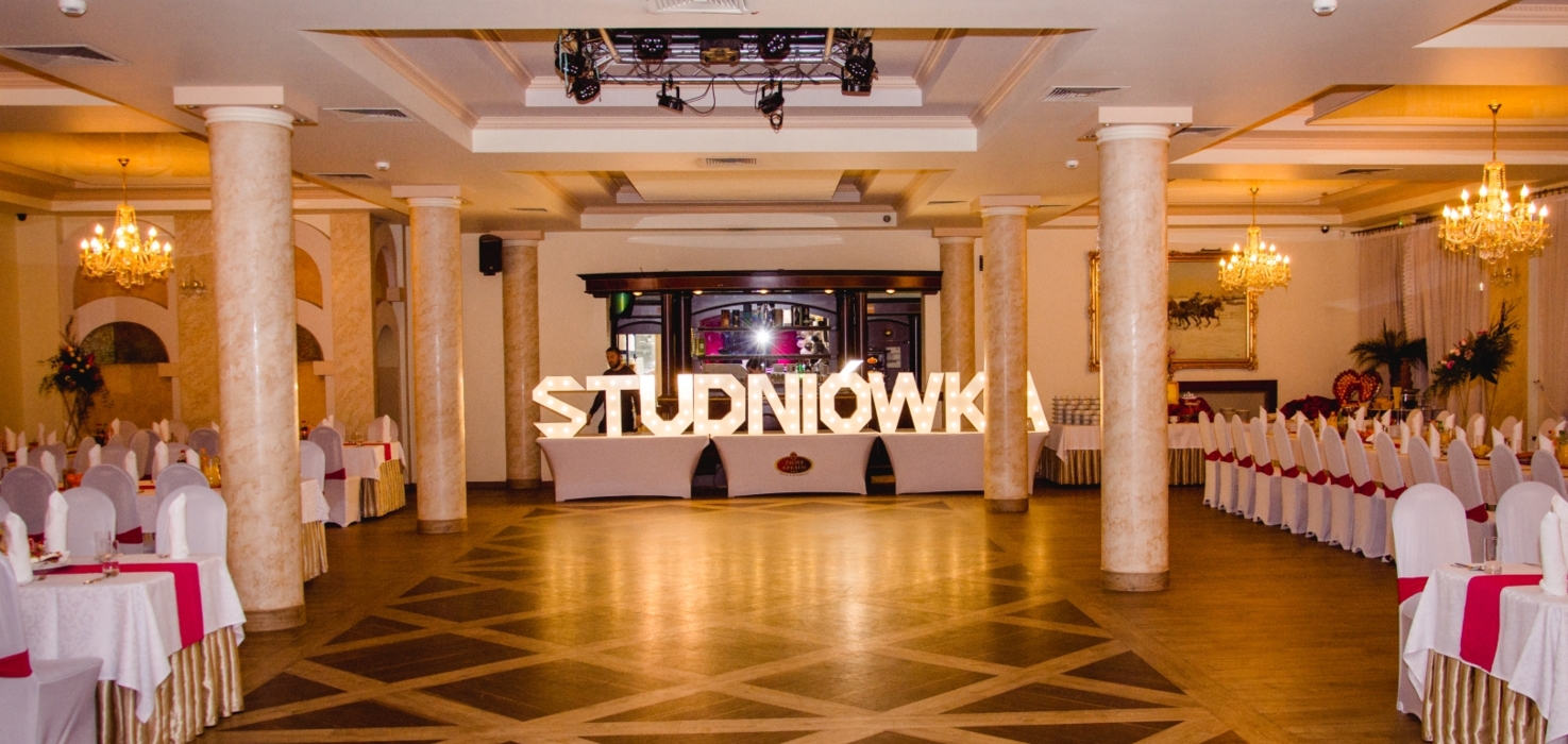 Elegancka sala balowa z jasnymi kolumnami, kryształowymi żyrandolami i stołami na bankiet z białymi pokrowcami i bordowymi akcentami, napis Studniówka