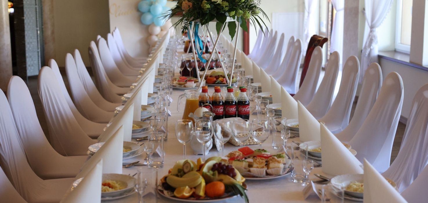 Długi stół z białym obrusem i pokrowcami na krzesła przygotowany na elegancką uroczystość z kwiatową dekoracją i napojami