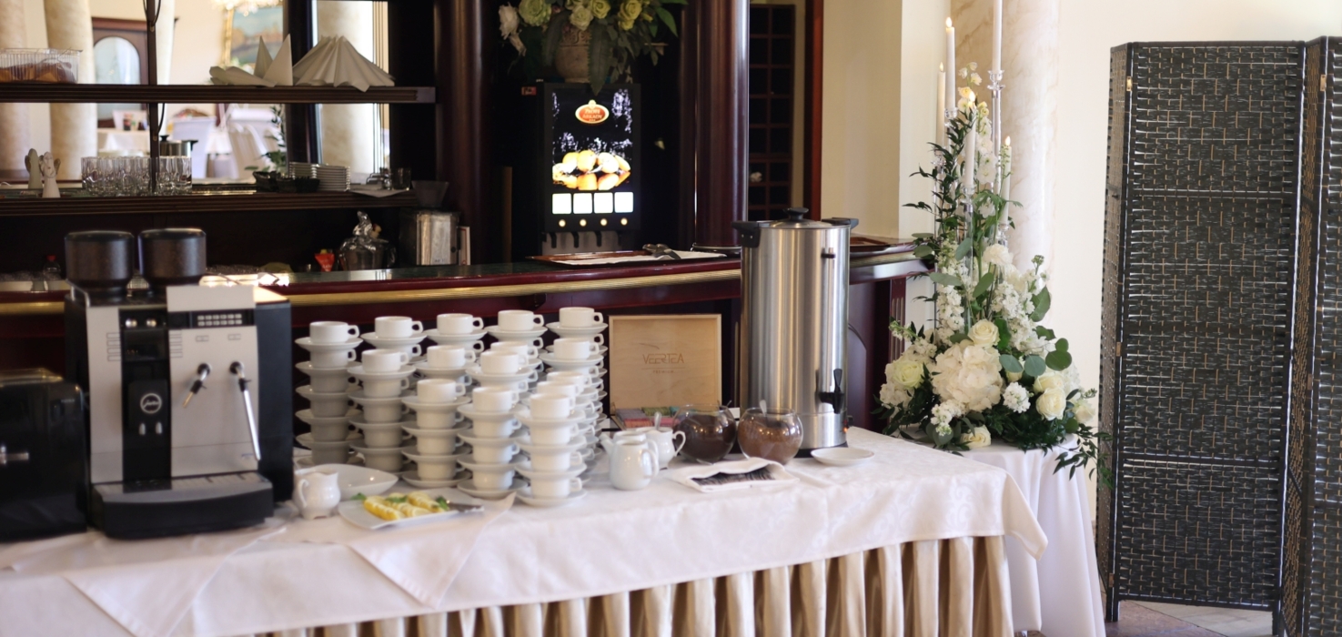 Elegancki bufet z kawą i herbatą, biały obrus, ekspres do kawy, stojak z filiżankami oraz dekoracje kwiatowe i świece.