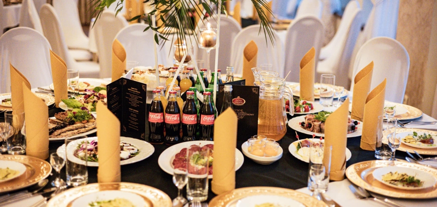 Elegancko nakryty okrągły stół bankietowy z złotymi talerzami, złotymi serwetkami i różnorodnymi przekąskami oraz napojami