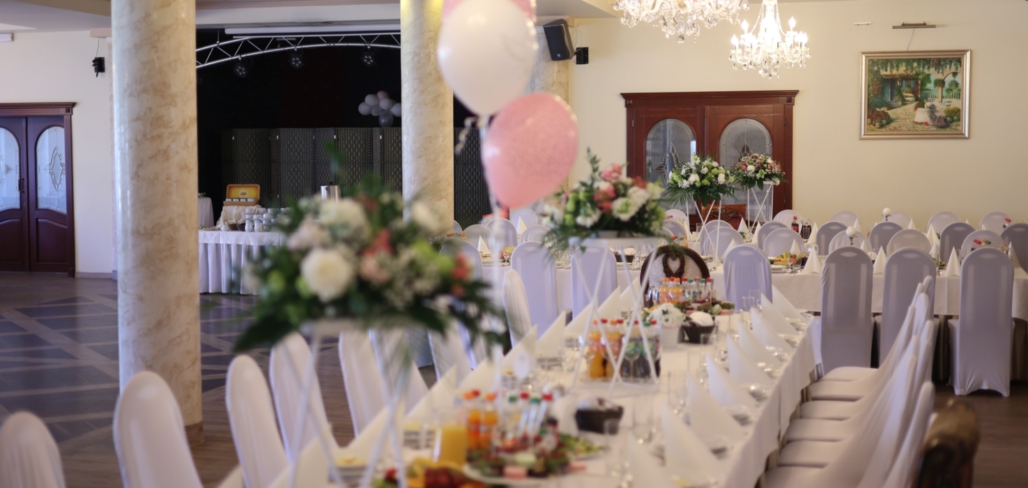 Elegancka sala bankietowa z długimi stołami w białych pokrowcach, kwiatowymi dekoracjami i różowymi balonami na przyjęcie