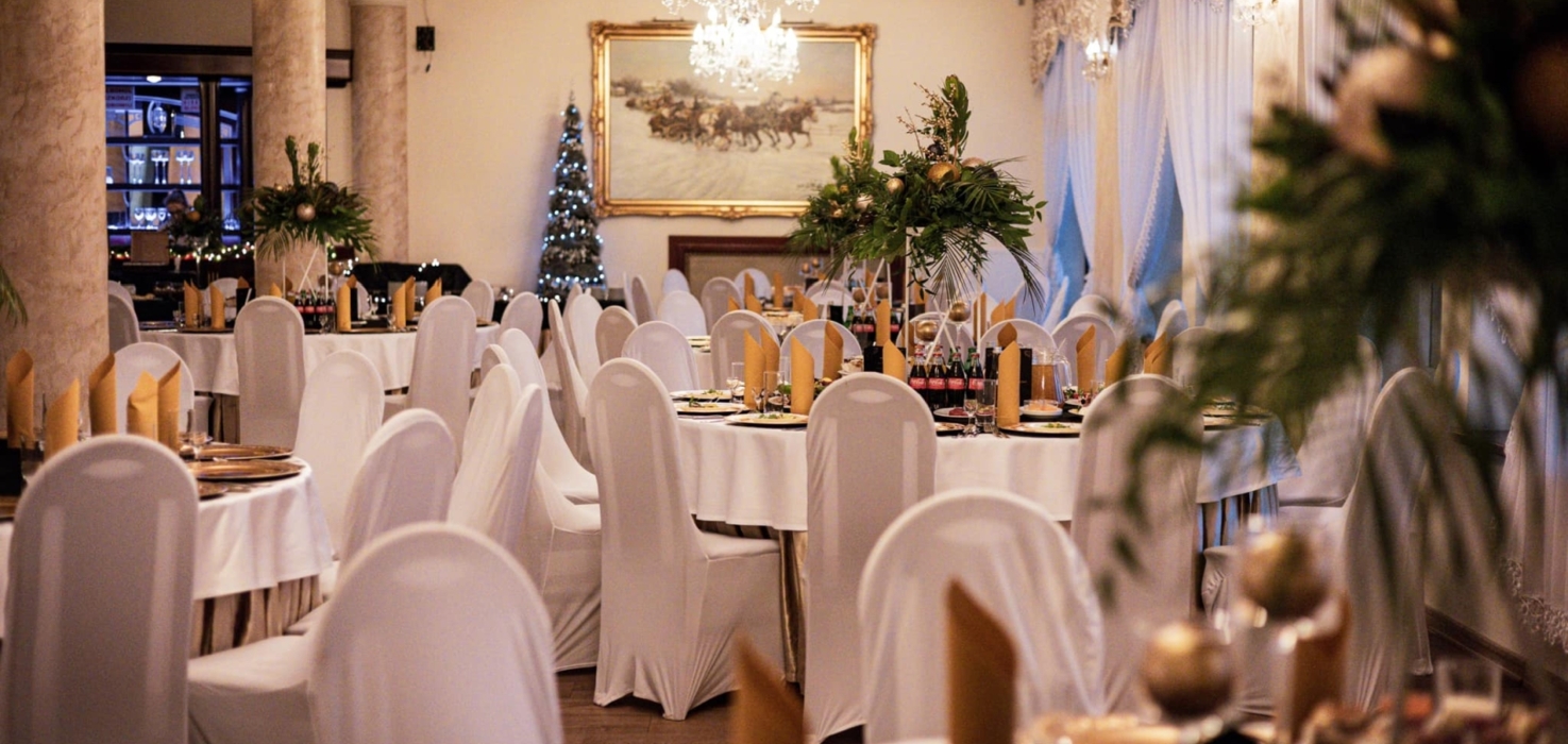 Elegancka sala bankietowa z białymi obrusami i pokrowcami, złotymi serwetkami, świąteczną choinką i kryształowymi żyrandolami.