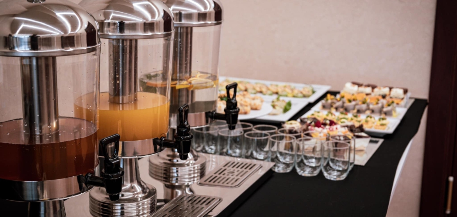 Bufet z trzema stalowymi dozownikami na soki i różnorodnymi przekąskami na talerzach, ustawionymi na czarnym obrusie