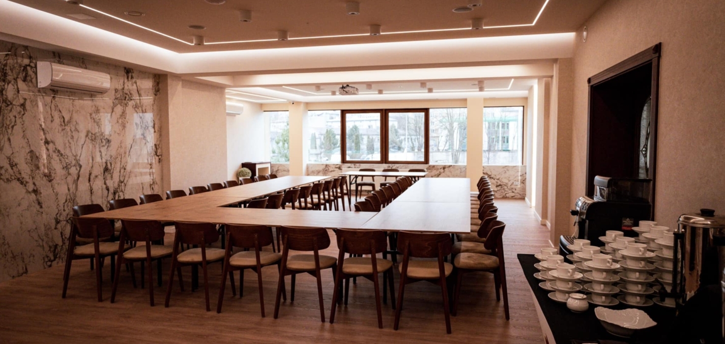 Nowoczesna sala konferencyjna z dużym stołem w kształcie litery U, drewnianymi krzesłami i kawowym bufetem przy ścianie