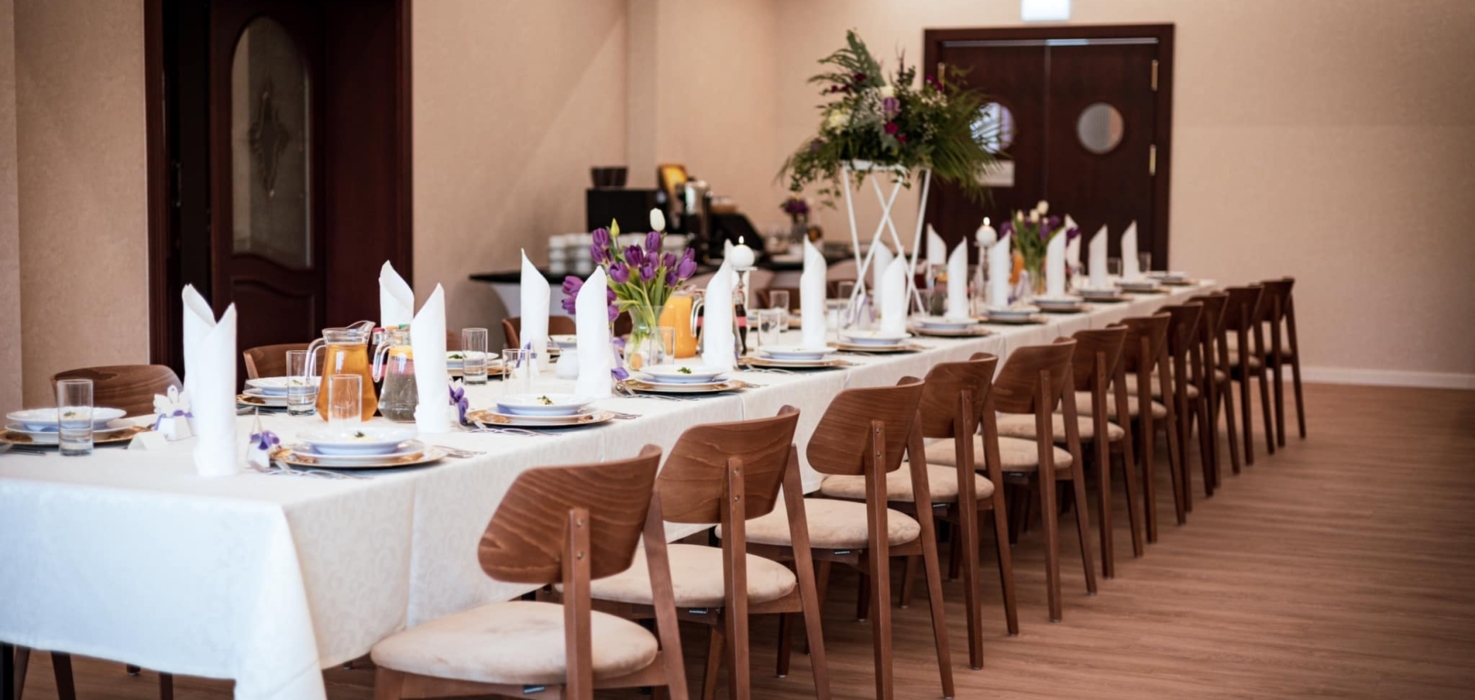 Długa sala bankietowa z elegancko nakrytym stołem, białe obrusy, fioletowe tulipany i drewniane krzesła na spotkania i uroczystości