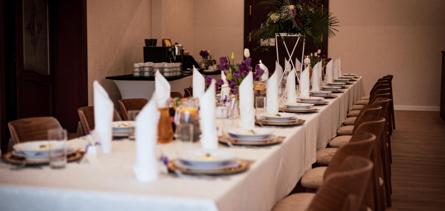 Długi stół bankietowy z białym obrusem, elegancko zastawiony talerzami, białymi serwetkami i bukietami kwiatów na kameralne przyjęcie