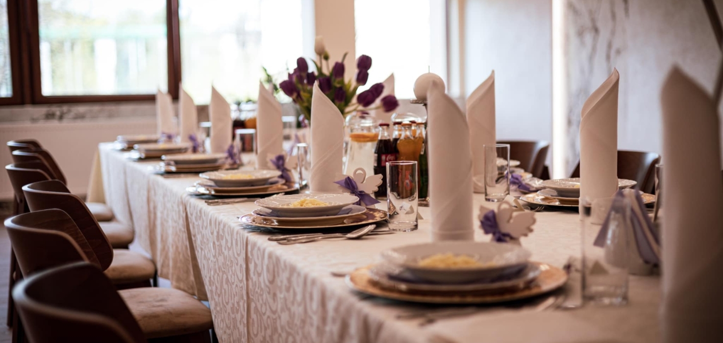 Elegancko nakryty stół bankietowy z białymi obrusami, serwetkami i kwiatową dekoracją w jasnym, przyjaznym wnętrzu.