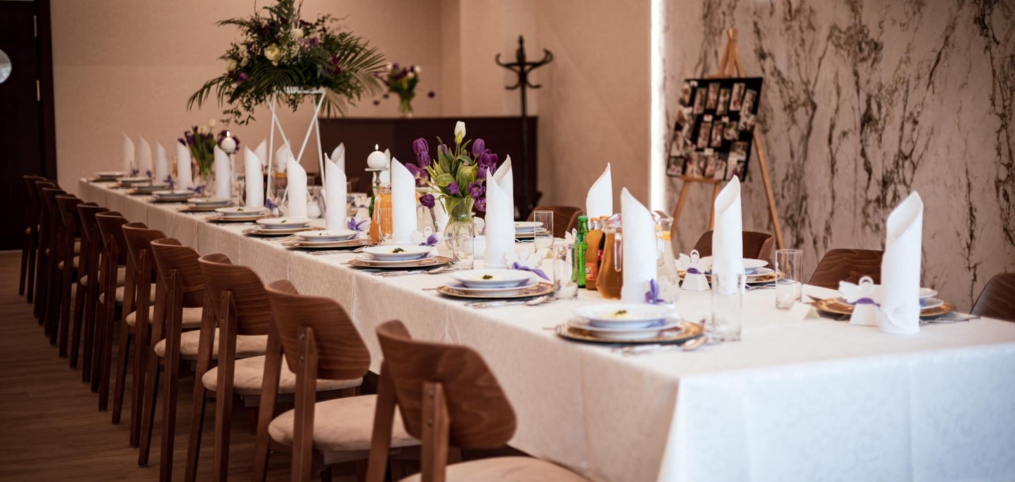 Elegancko nakryty długi stół w sali bankietowej, z białymi serwetkami, kwiatowymi dekoracjami i drewnianymi krzesłami