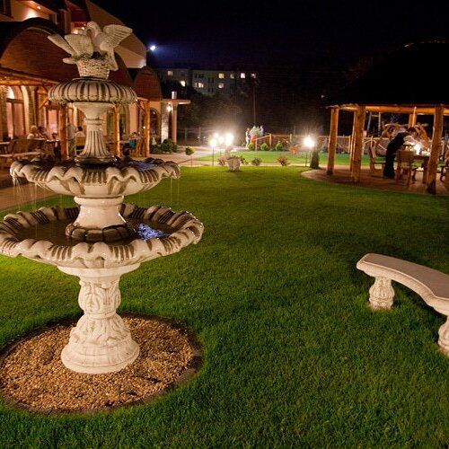Oświetlony nocą ogród hotelowy z ozdobną fontanną, ławką na trawniku i zadaszonym miejscem do relaksu