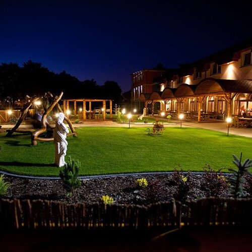 Oświetlony na noc hotel z rozległym zielonym ogrodem, rzeźbą i drewnianą huśtawką, tworzący spokojną, przyjazną atmosferę.