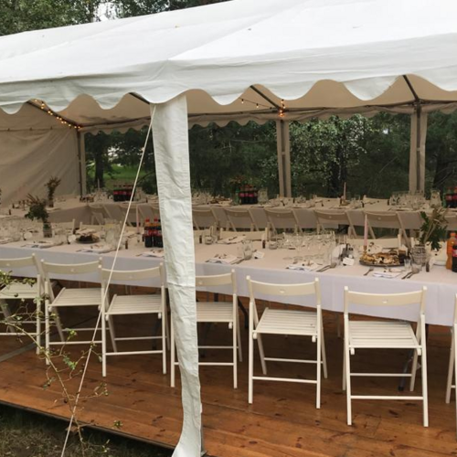 Przyjęcie na zewnątrz pod białym namiotem z długimi stołami udekorowanymi białymi obrusami, kwiatami i napojami.
