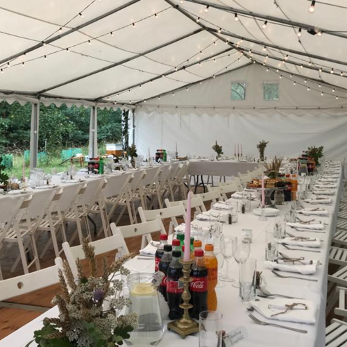 Przyjęcie w dużym namiocie z długimi stołami, białymi krzesłami, dekoracjami i napojami, w otoczeniu zieleni