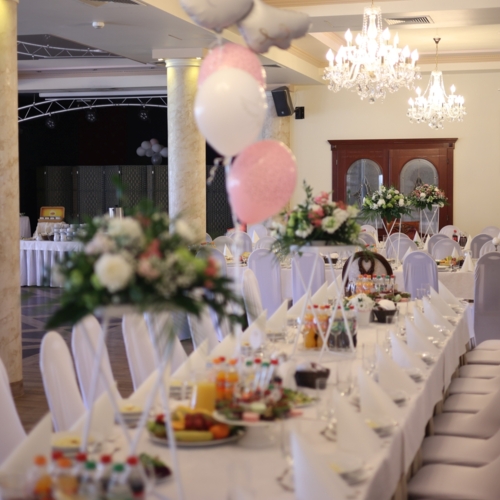 Elegancka sala bankietowa z długimi stołami w białych pokrowcach, kwiatowymi dekoracjami i różowymi balonami na przyjęcie